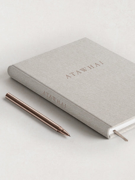The front cover of the Atawhai Planner for neurodivergents and people with ADHD, featuring a soft grey linen hard coder, embossed rose gold lettering and matching ribbon marker. A rose gold metal pen is placed delicately next to the journal.