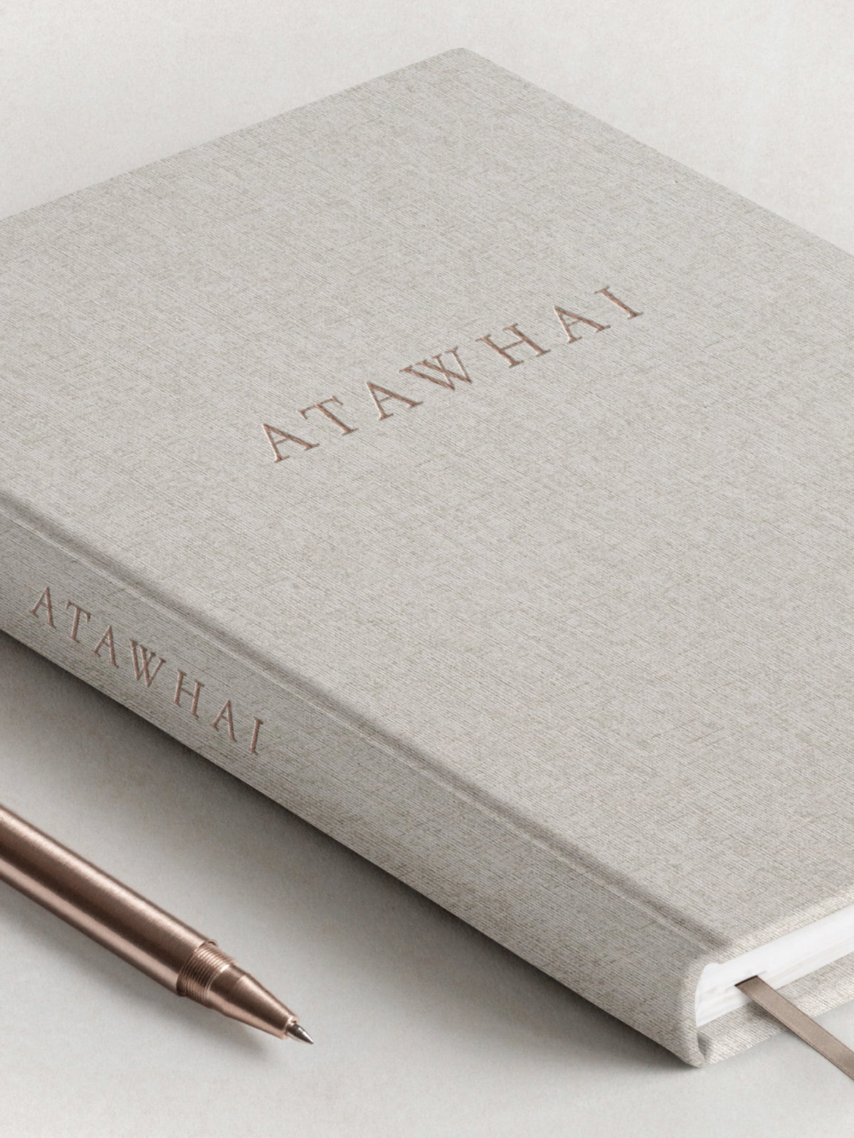 The front cover of the Atawhai Planner for neurodivergents and people with ADHD, featuring a soft grey linen hard coder, embossed rose gold lettering and matching ribbon marker. A rose gold metal pen is placed delicately next to the journal.
