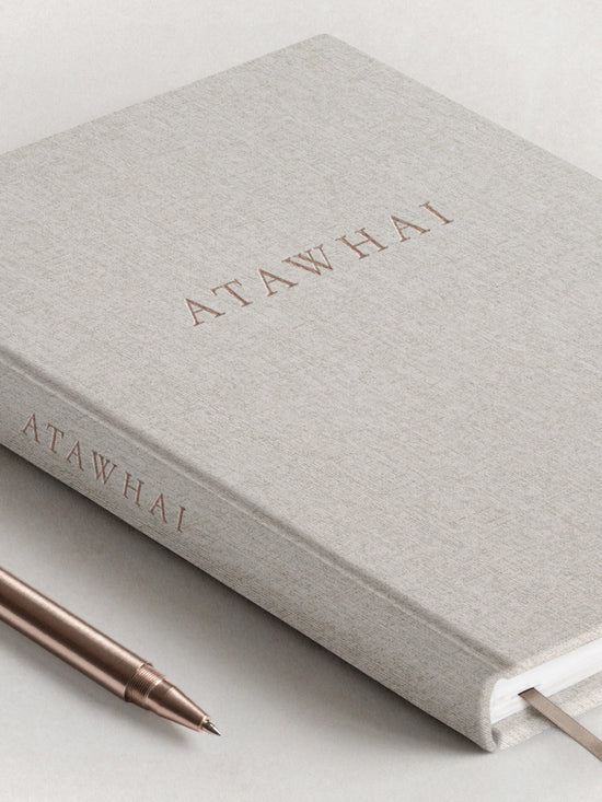The front cover of the Atawhai Planner for neurodivergents and people with ADHD, featuring a soft grey linen hard coder, embossed rose gold lettering and matching ribbon marker. A rose gold metal pen is placed delicately next to the journal.