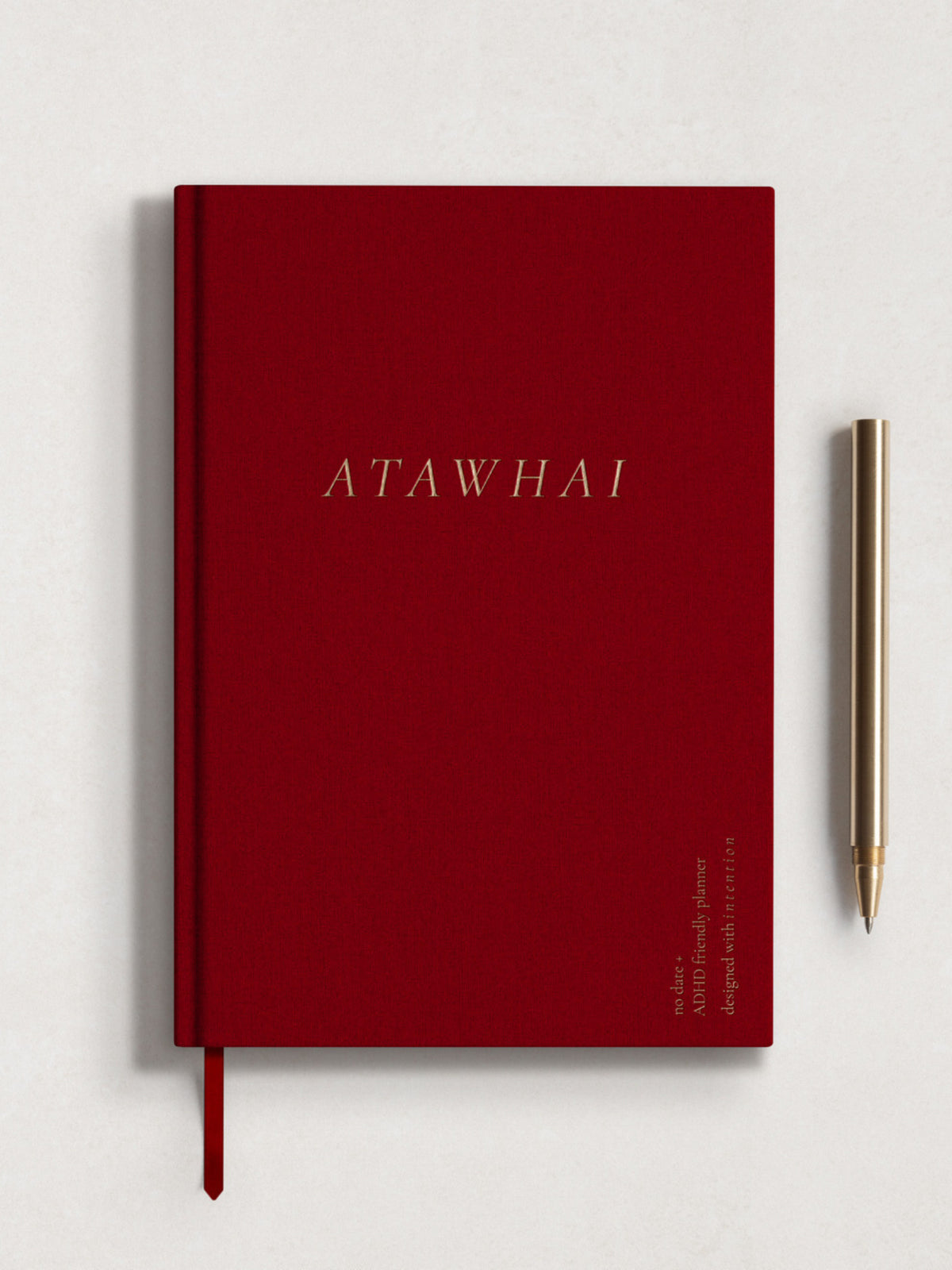 The front cover of the Atawhai Planner for neurodivergents and people with ADHD, featuring a soft red linen hard cover, embossed  gold lettering and matching ribbon marker. A gold metal pen is placed delicately next to the journal.